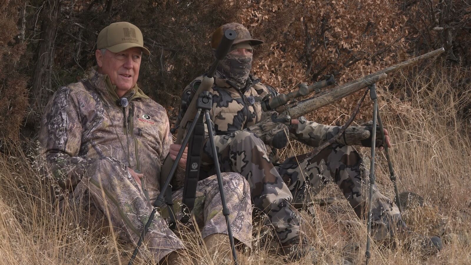 A guide and shooter seated side by side in tall winter grass, rifle steadied on a tripod between them