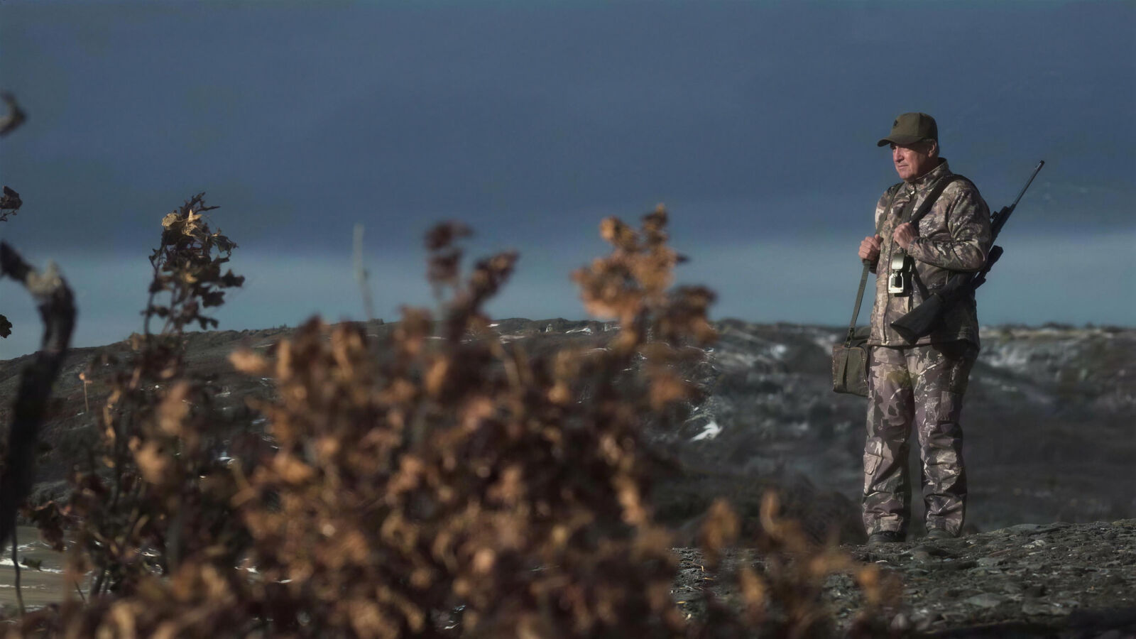 A hunter in camouflage glassing a snow-patched valley from a rocky ridge under a heavy storm sky, rifle slung on his shoulder