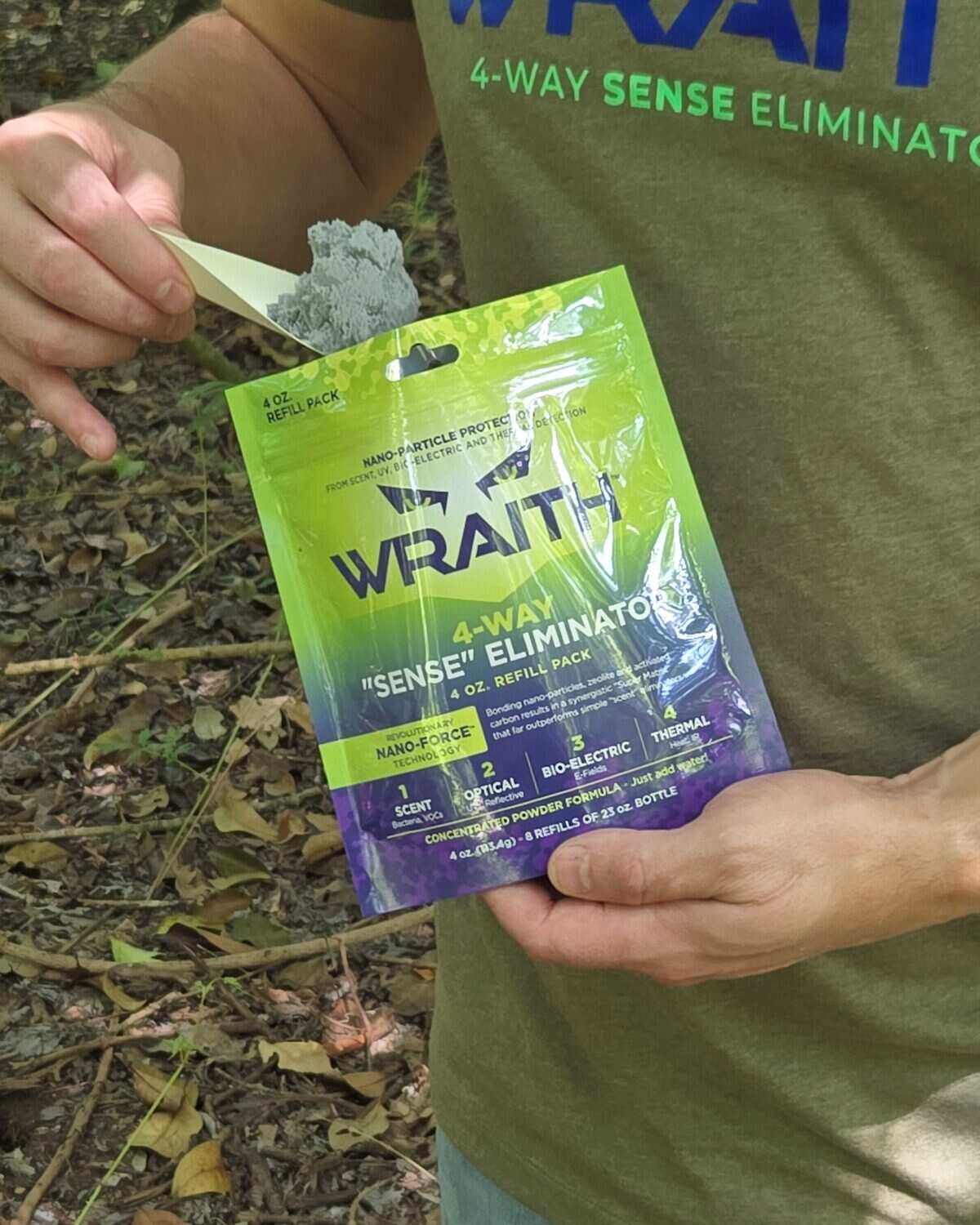 A hunter in a WRAITH t-shirt holding the 4 oz refill pack in one hand and a folded paper scoop heaped with gray powder in the other, forest floor behind